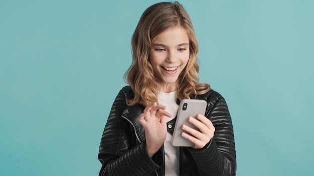 Beautiful blond teenager girl chatting with friends on smartphone looking happy isolated on colorful background