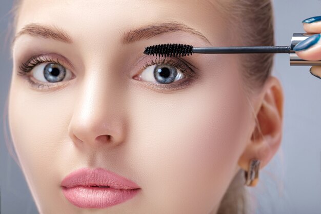 beautiful blond woman applying makeup on face on gray background. perfect makeup for blue eyes. the brush of mascara