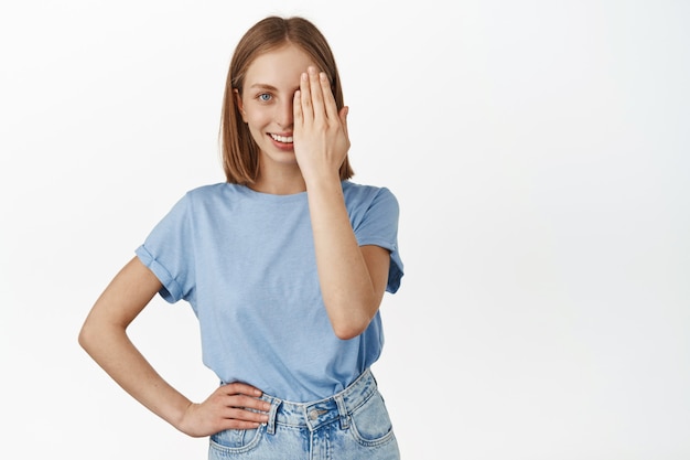 美しいブロンドの女性が顔の半分を覆い、片目で見て自信を持って笑顔で、効果の前後に、白い壁に青いTシャツを着て立っています