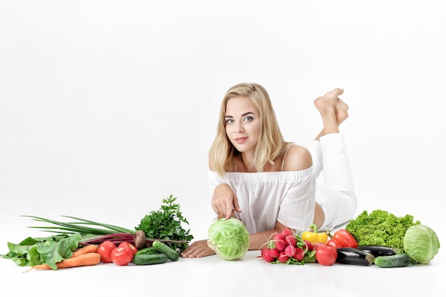 Фото Красивая белокурая женщина в белой одежде и много свежих овощей на белом фоне