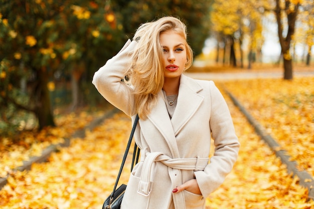 Beautiful blond woman in a trendy coat with a handbag walking in an autumn park of yellow foliage