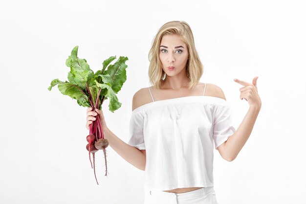 Beautiful blond woman in a white blouse is pointing at beetroot with green leaves on a white background. Health and vitamins