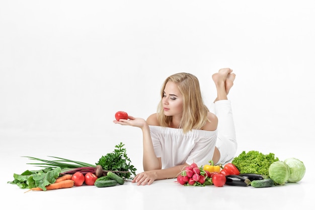 Красивая блондинка женщина в белой одежде и много свежих овощей на белом фоне. Девушка держит помидор