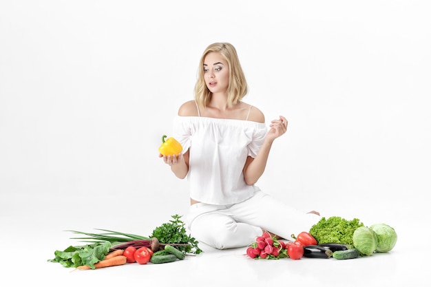 Beautiful blond woman in white clothes and lots of fresh vegetables on a white background. Girl holds bell pepper