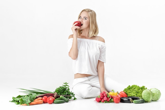 Bella donna bionda in abiti bianchi e un sacco di verdure fresche su uno sfondo bianco. la ragazza sta mangiando la nettarina