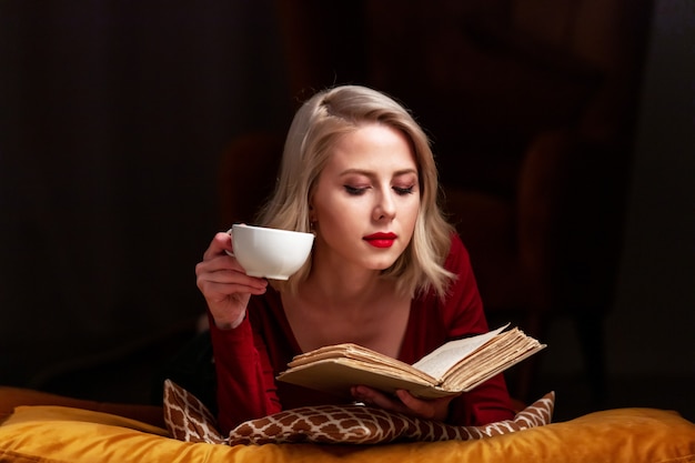 Beautiful blond woman with book and cup lies on pillows