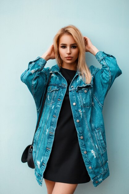 Beautiful blond woman with handbag in blue denim clothes posing near blue wall