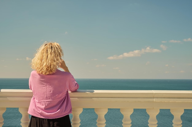 ピンクの t シャツを着た美しい金髪の大人の女性が立って、バルコニーから海の景色を楽しみますテキスト用の背面ビュー スペース