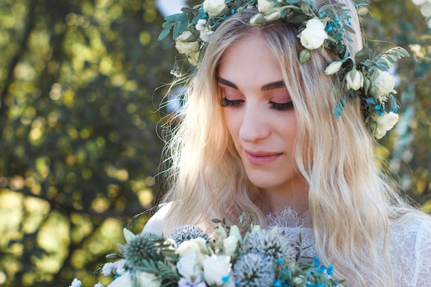 Beautiful blonde bride portrait with flower bouquet and wreath on her head in white wedding dress