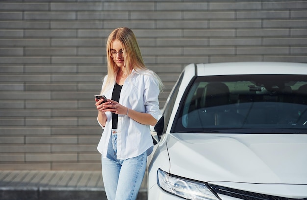 Foto una bella bionda in abiti casuali con il telefono vicino a un'auto bianca è all'aperto durante il giorno soleggiato