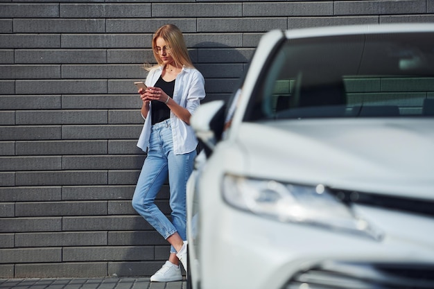 Beautiful blonde in casual clothes with phone near white car is outdoors at sunny daytime