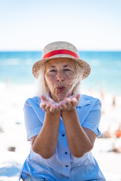 Photo beautiful blonde caucasian woman in a straw hat and striped shirt blowing air kiss sitting by the sea in summer