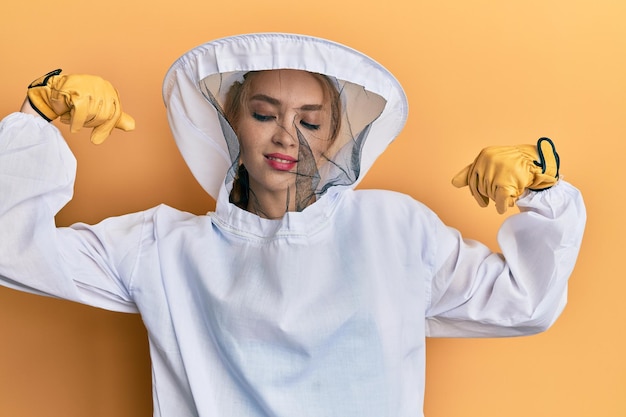 Beautiful blonde caucasian woman wearing protective beekeeper uniform showing arms muscles smiling proud. fitness concept.