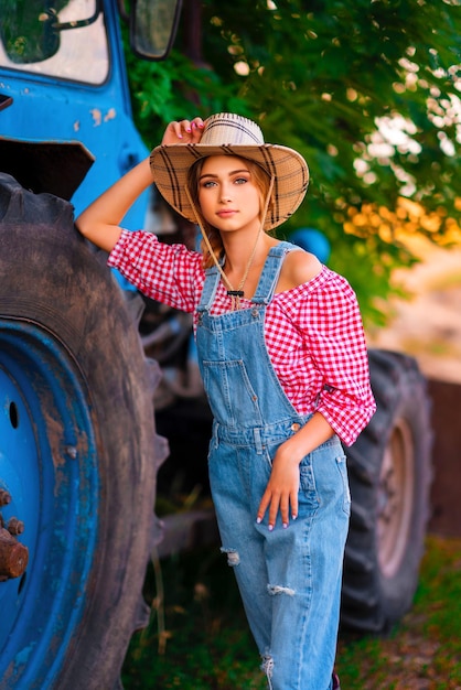 Beautiful blonde cowgirl in straw hat and denim overall standing near blue tractor.