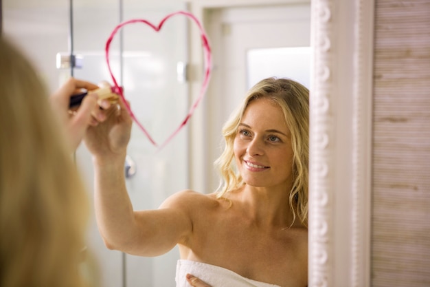 Beautiful blonde drawing big heart on mirror in the bathroom at home