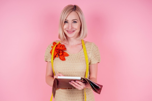 La bella sarta femminile bionda scrive il modello della curva sarto crea una collezione abiti cuce vestiti macchina da cucire in officina. giovane donna vestiti firmati note idee sfondo rosa studio