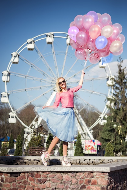 Bella ragazza bionda in un parco divertimenti, ruote panoramiche con un grosso mazzo di palloncini in mano. colori rosa e blu.