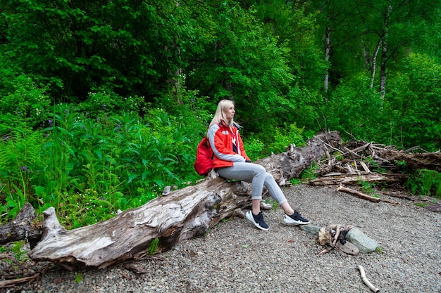 Beautiful blonde girl in a autumn jacket with a red backpack sits on a fallen tree in a forest or park in nature walking and traveling in fresh air loneliness and relaxation from bustle of the city