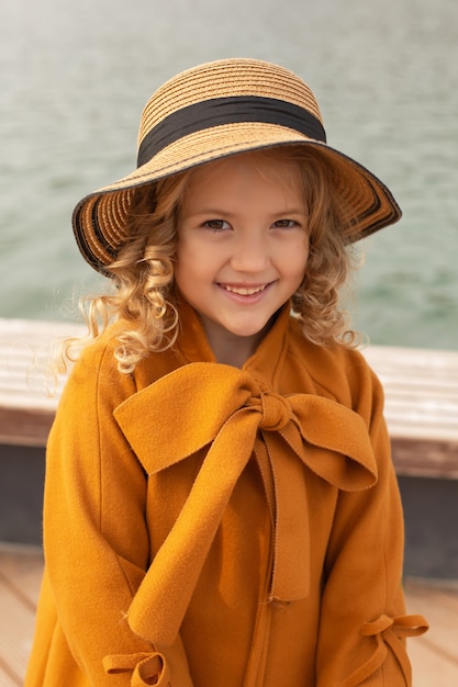 街の湖の自然の桟橋に帽子をかぶったベージュのコートを着た美しいブロンドの女の子