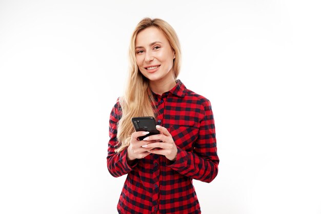 Beautiful blonde girl in casual looks at the smartphone screen and smiles isolated on white studio background