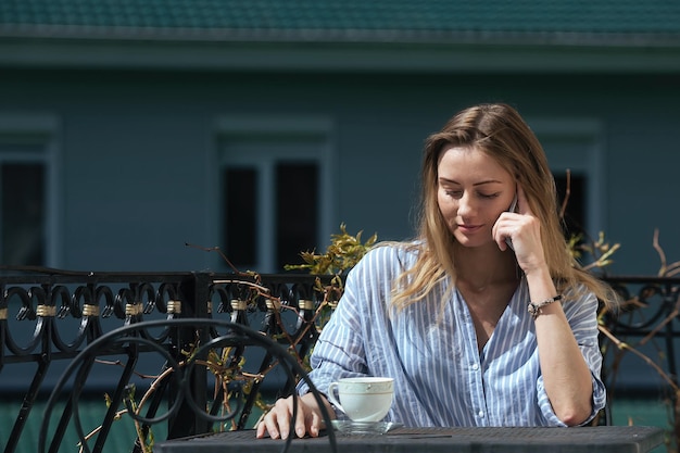 beautiful blonde girl drinks coffee at a table on the balcony of her house and talks on the phone