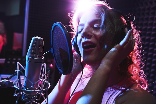 Bella ragazza bionda che canta emotivamente una canzone in studio di registrazione con microfono professionale e cuffie crea un nuovo artista vocale dell'album della traccia in rosa blu luce al neon closeup faccia