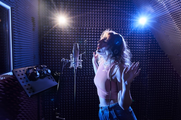 プロのマイクとヘッドフォンを備えたレコーディングスタジオで感情的に歌を歌っている美しいブロンドの女の子は、ピンクブルーのネオンライトで新しいトラックアルバムのボーカルアーティストを作成します