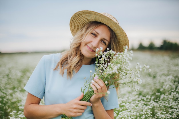 ヒナギクの分野で美しいブロンドの女の子。花のフィールドに青いドレスの女。ヒナギクの花束を持つ少女。村の夏の入札写真。野の花。麦わら帽子の少女