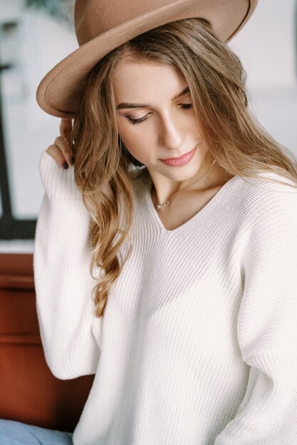 A beautiful blonde girl in a hat sits on a brown sofa