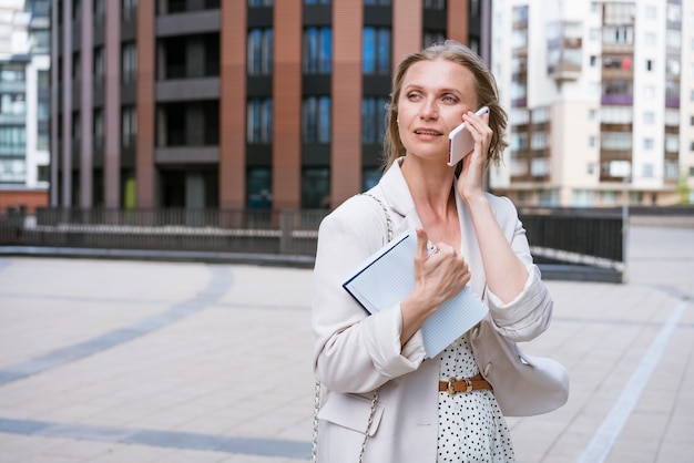 A beautiful blonde girl holds a smartphone and a notebook in her hands business woman uses a mobile ...