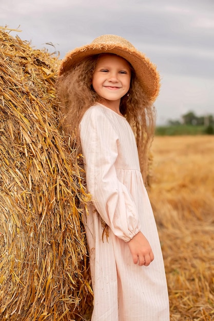 a beautiful blonde girl in a linen dress and a straw hat stands by a haystack in the field