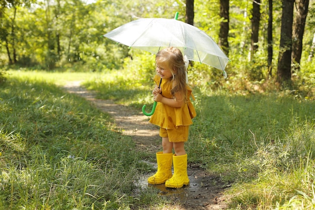 透明な傘とリネンのスーツを着た美しいブロンドの女の子が公園の雨の中に立っています