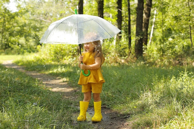 透明な傘とリネンのスーツを着た美しいブロンドの女の子が公園の雨の中に立っています