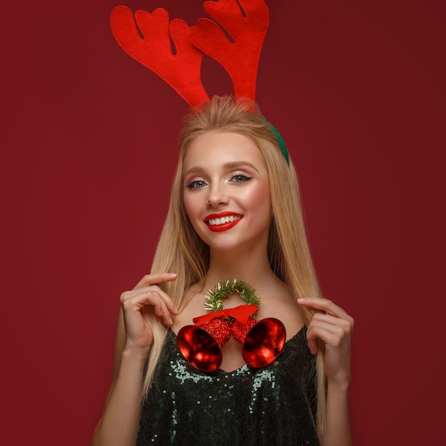 Bella ragazza bionda in un'immagine di capodanno con campane di natale al collo e corna di cervo sulla testa. fronte di bellezza con trucco festivo. foto scattata in studio.