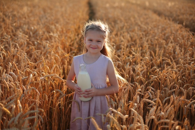 a beautiful blonde girl in a pink linen dress is sitting in a wheat field with a bottle of milk