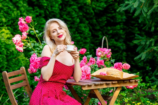 Bella ragazza bionda in vestito rosso che beve un caffè in un giardino