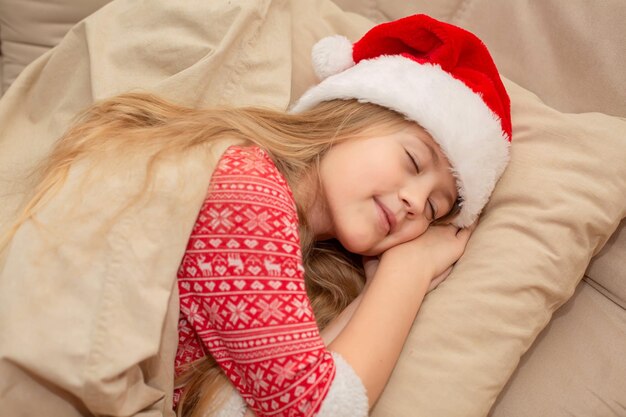 Photo a beautiful blonde girl in a santa claus hat and new year's pajamas is sleeping in bed