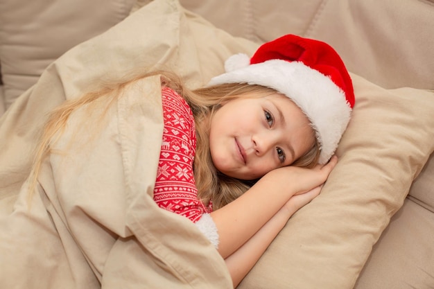 a beautiful blonde girl in a Santa Claus hat and New Year's pajamas is sleeping in bed