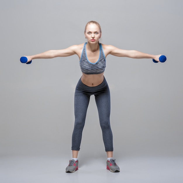 beautiful blonde girl in sportswear does exercises with dumbbells on a grey background