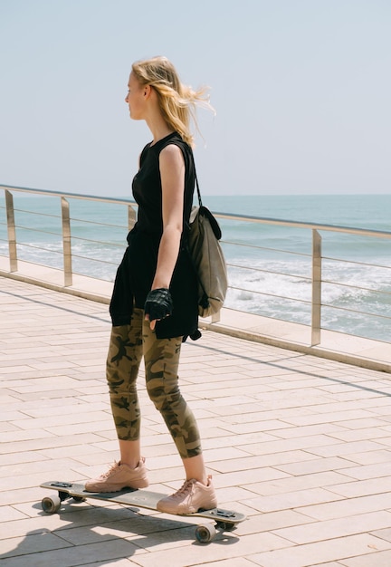 Красивая блондинка с рюкзаком на скейтборде в жаркий летний день на берегу моря. Ветер развевает волосы. Наслаждение свободой