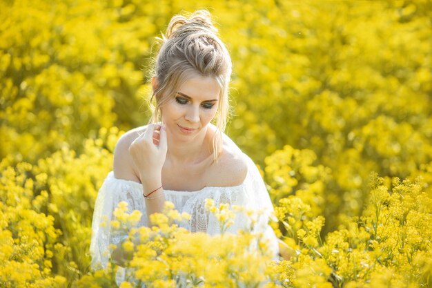 Foto bella ragazza bionda con i capelli lunghi in un campo di colza in un abito bianco in un campo di fiori gialli che sbocciano in un campo di colza al tramonto o all'alba