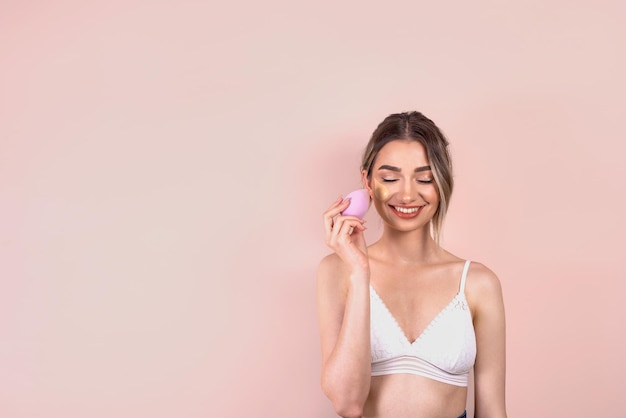 Beautiful blonde girl with natural makeup on light pink background applying foundation on her face with a beauty blender copy space