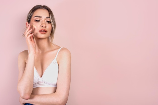 Beautiful blonde girl with natural makeup on light pink background blending foundation on her face copy space