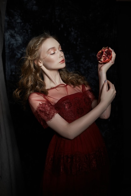 Bella ragazza bionda con il frutto del melograno nelle sue mani. ritratto di primavera di una ragazza in un vestito rosso che rompe un melograno, succo che scorre lungo le sue mani