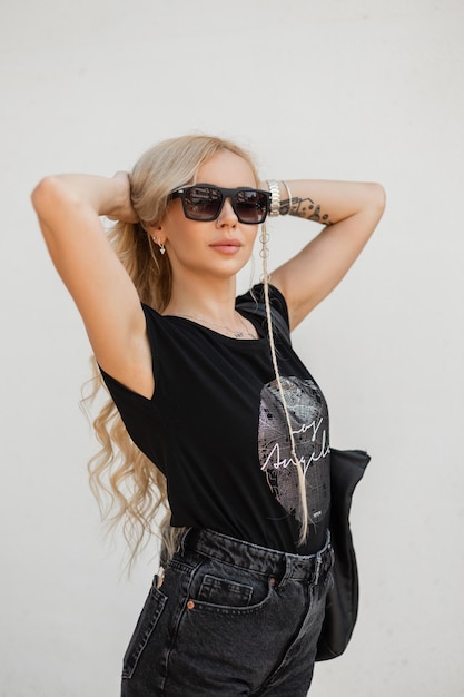 Beautiful blonde girl with stylish modern sunglasses in fashion black summer clothes with denim shorts a Tshirt and a trendy bag on a white background on the street