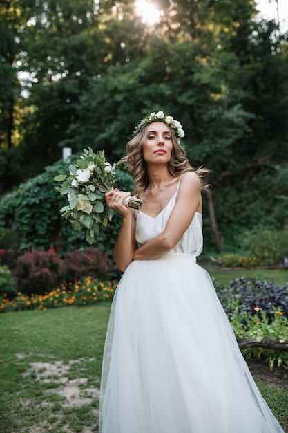 Красивая блондинка счастливая невеста в элегантном белом платье в венке с букетом на открытом воздухе
