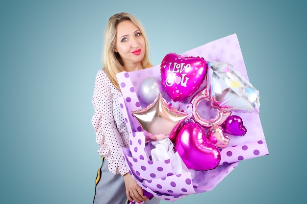 A beautiful blonde holds in her hands a bouquet of balloons in the shape of hearts on which the inscription I love you