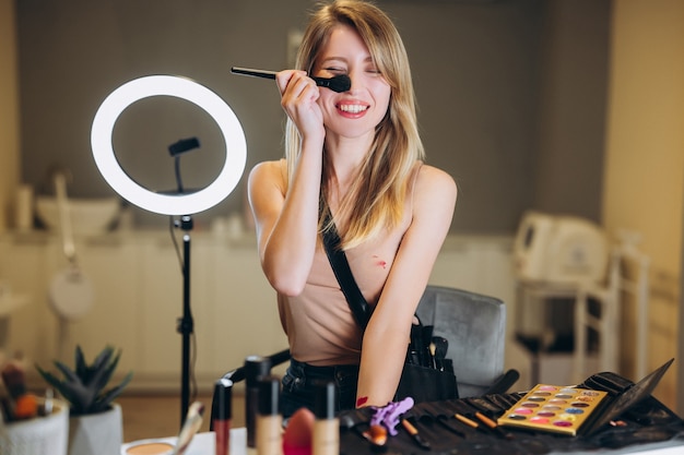 The beautiful blonde is laughing and closes one eye with a brush for makeup. Young beautiful woman holding makeup brush near her nose.