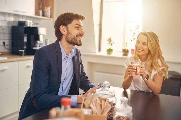 Beautiful blonde lady looking at bearded gentleman and smiling while sitting at the table and holding cup of hot drink