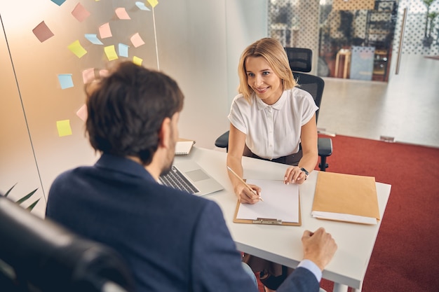 Beautiful blonde lady looking at man and smiling while sitting at the table and writing on clipboard